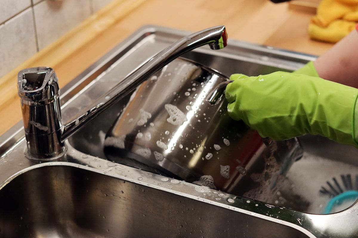 Washing dishes in a stainless steel sink with green rubber gloves.